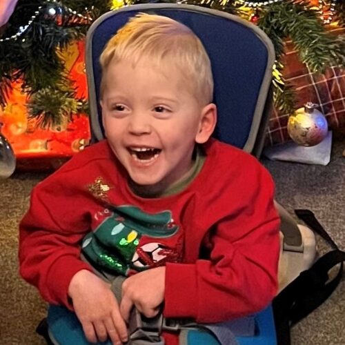 A three-year old boy sits in a go-to seat smiling. He is wearing a Christmas jumper