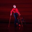 A man in a red shirt, black trousers and red shoes, poses holds a air of crutches with his wheelchair in the background