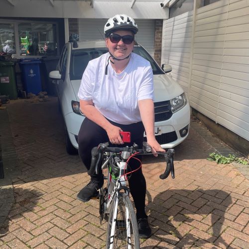 A woman wearing a white t-shirt, black leggings and a helmet sits in front of a driveway on her bike, smiling.