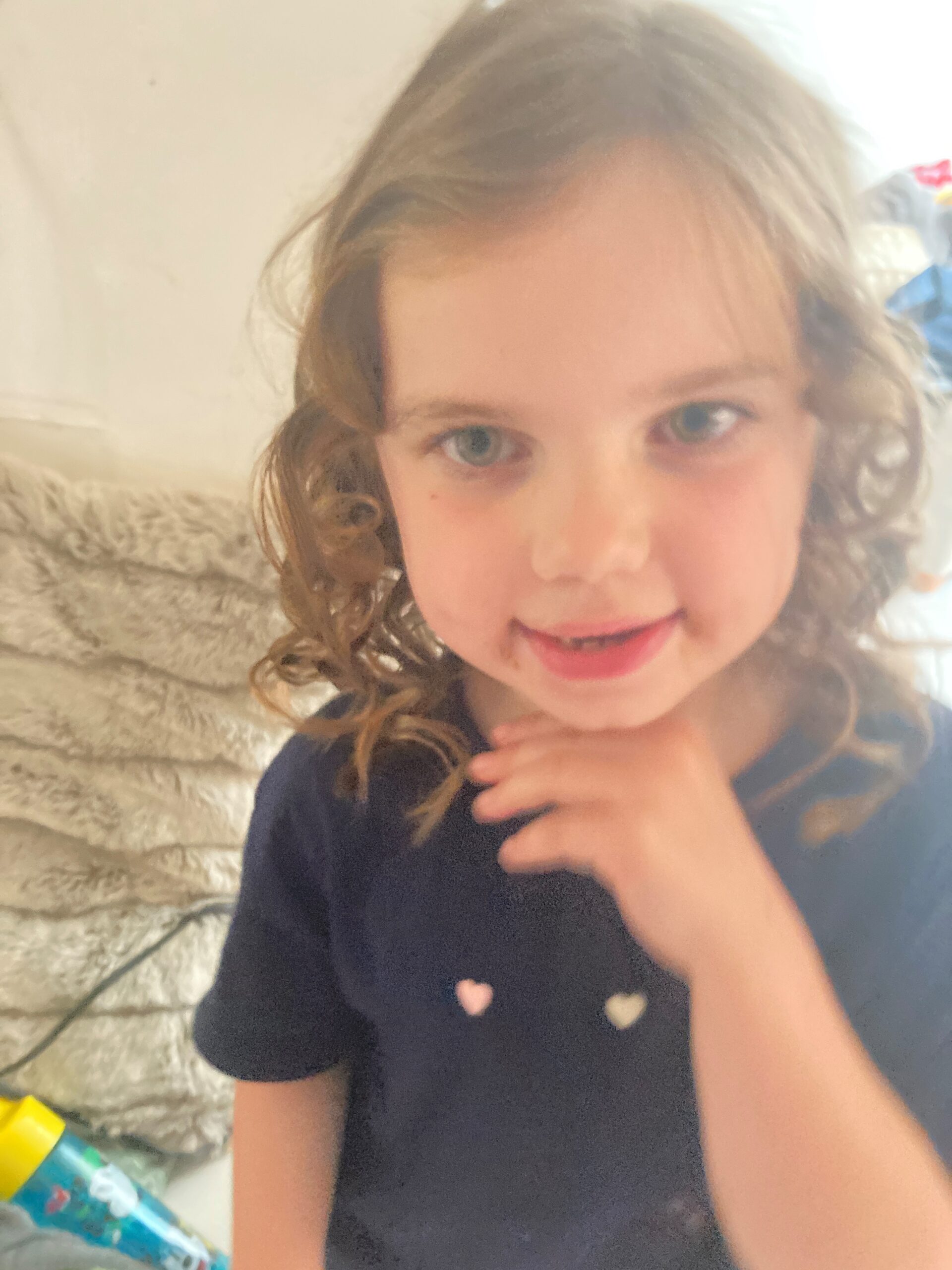 A little girl with curly hair holds her hand under her chin and is smiling 