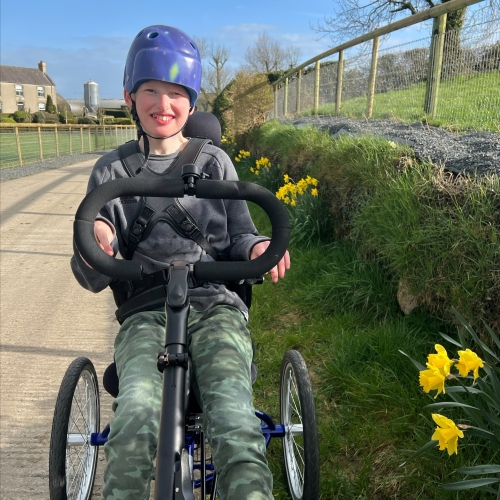 A teenage boy is smiling at the camera while sitting on an adapted trike. He is wearing a blue coat and black joggers